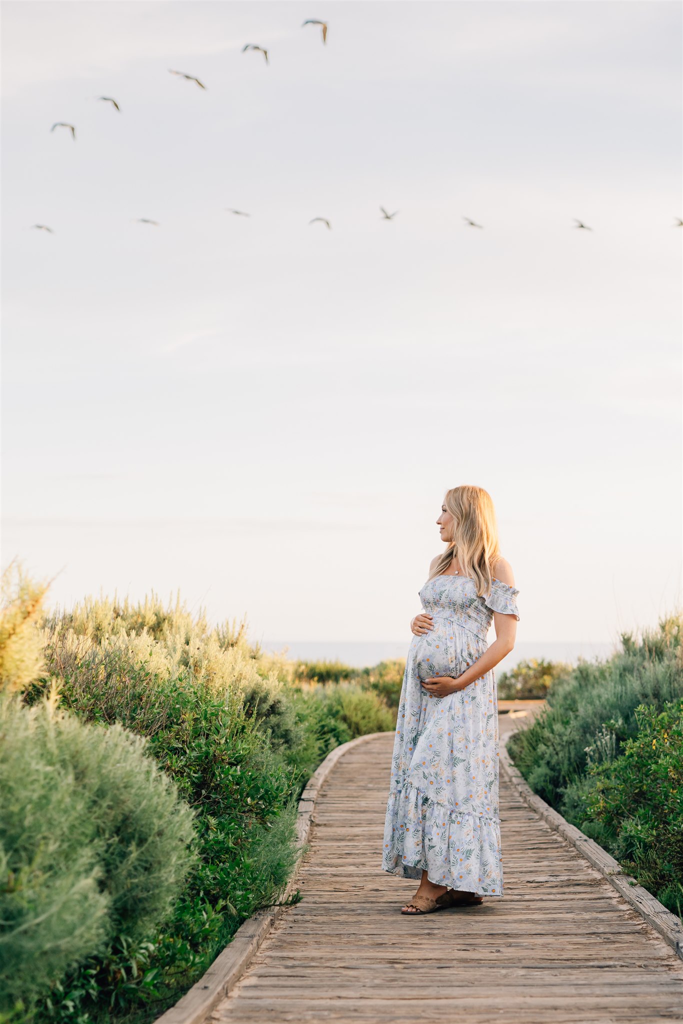 A happy mom to be walks on a beach boardwalk at sunset in a blue maternity dress