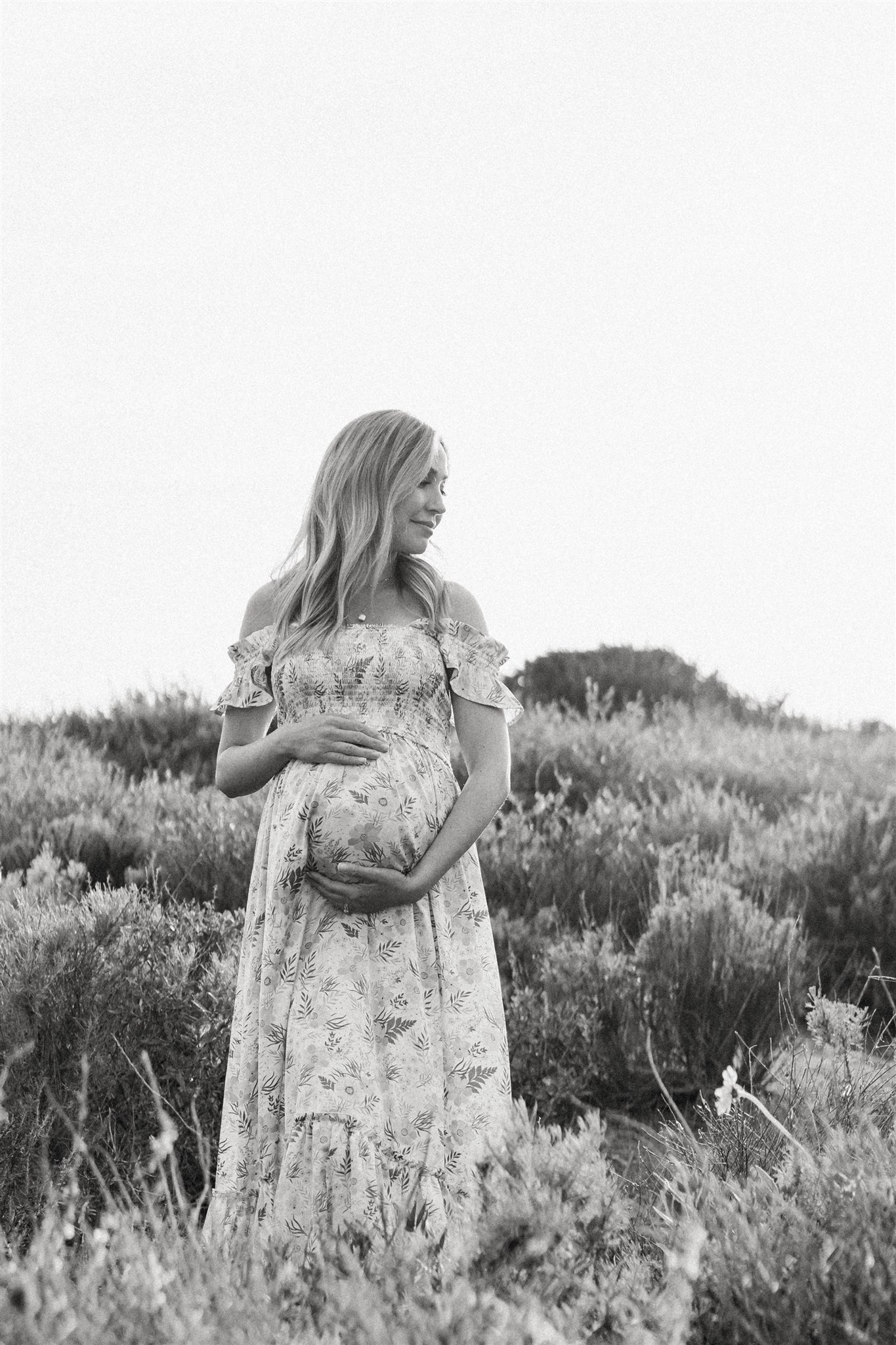 A mom to be stands on a dune smiling down her shoulder and holding her bump during Family Day Trips in Orange County