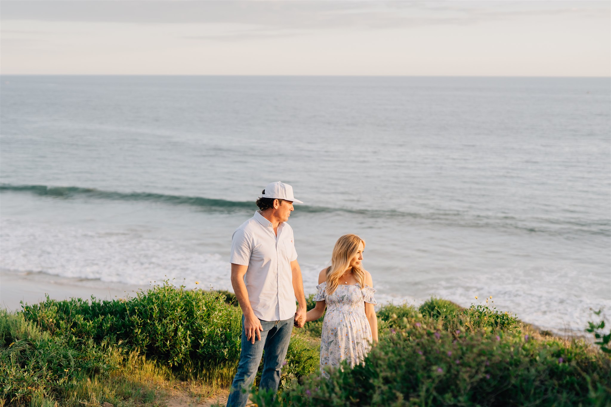 A pregnant mom and dad walk on a beach dune watching the sunset during Family Day Trips in Orange County