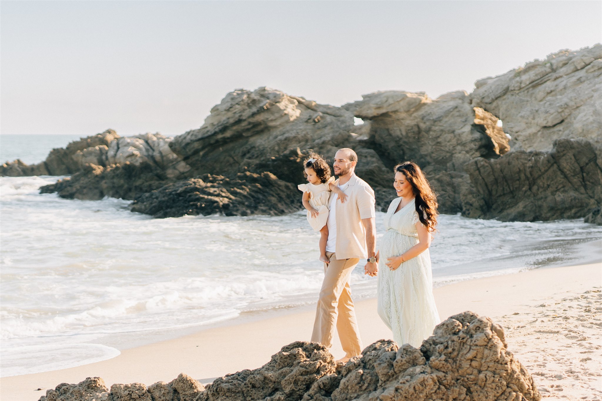 A happy mom and dad walk on a beach at sunset with their toddler daughter in dad's arms