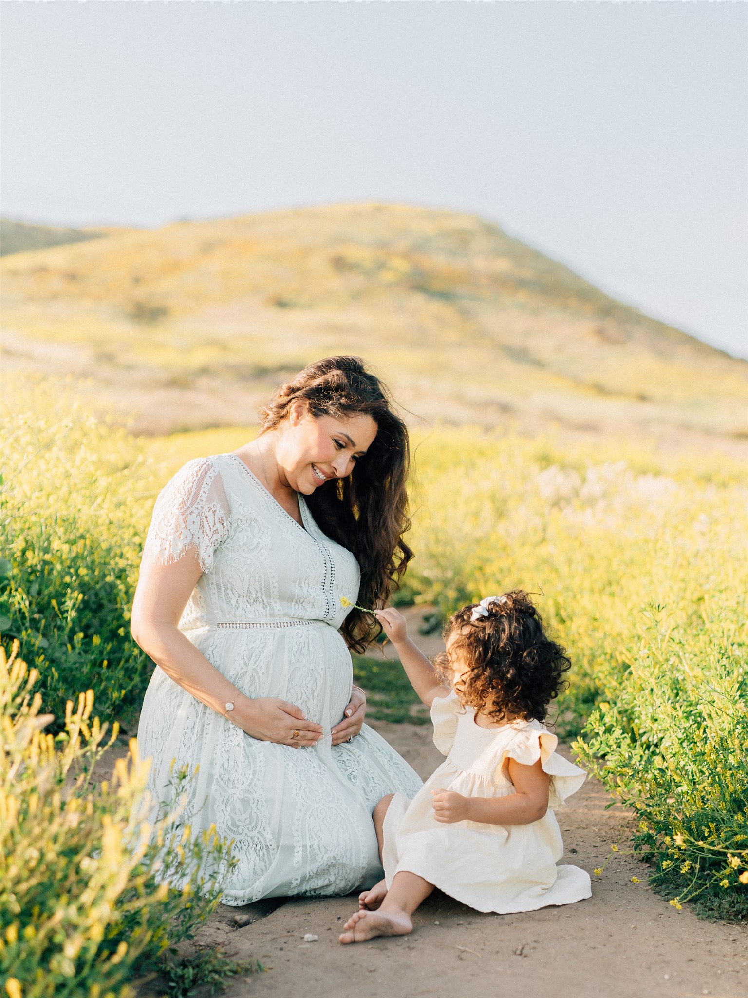 A pregnant mom plays with her toddler daughter picking yellow wildflowers in a park trail at sunset