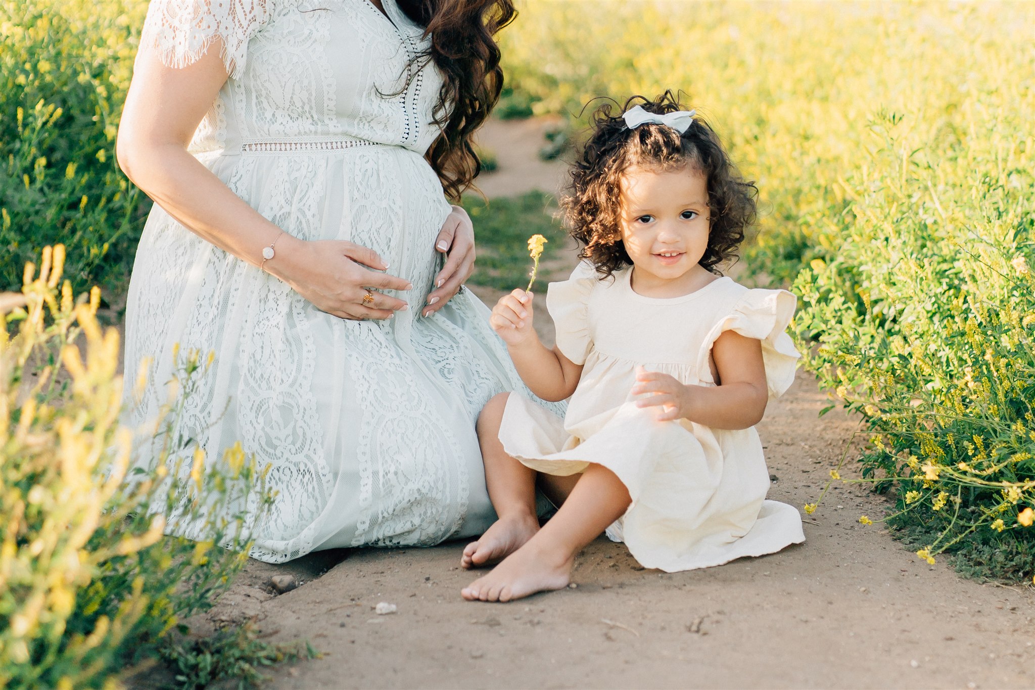 A toddler girl sits in a park trail surrounded by wildflowers with her pregnant mom before visiting Preschools in Pacific Palisades