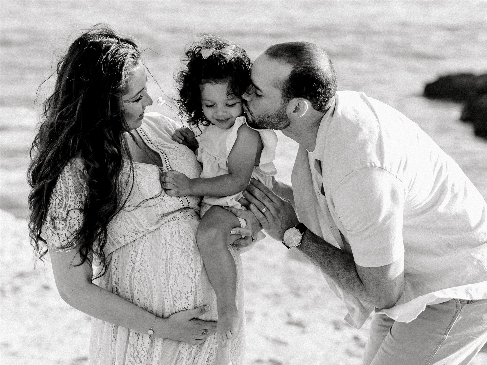 A dad kisses his toddler daughter sitting on mom's pregnant bump on a beach in black and white before visiting Preschools in Pacific Palisades